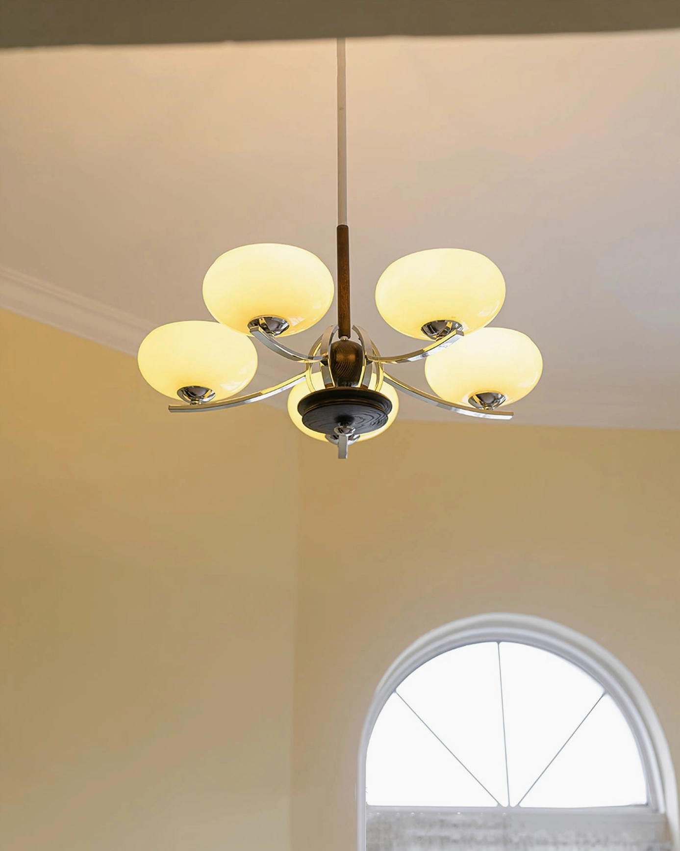 Five-light mid-century modern glass chandelier photographed from low upward angle in indoor space with arched window and warm vintage French country styling