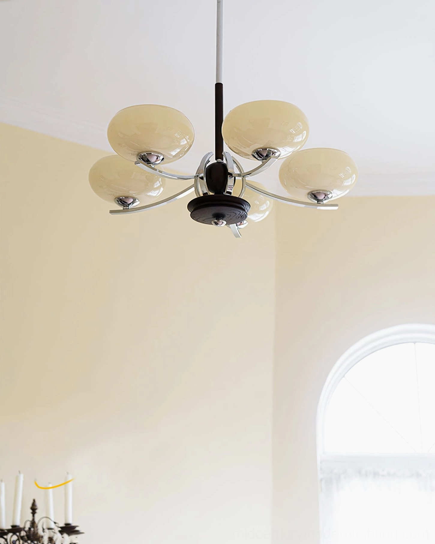 Five-light glass chandelier with dark wood central rod, curved chrome metal arms and cream-colored glass shades against yellow wall