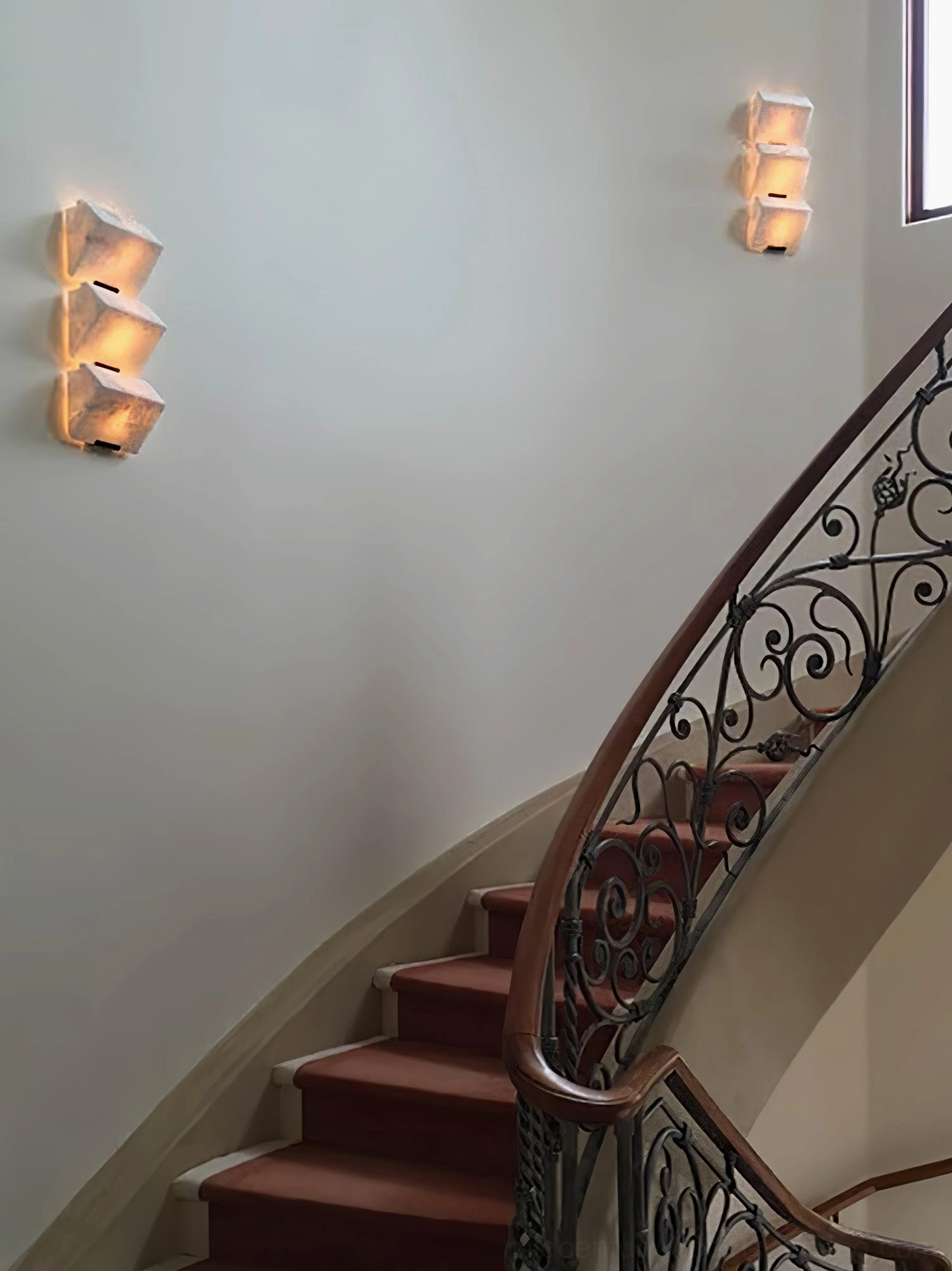 Pair of compact alabaster wall sconces installed on hallway near staircase with wrought iron railing, classic luxury home accent