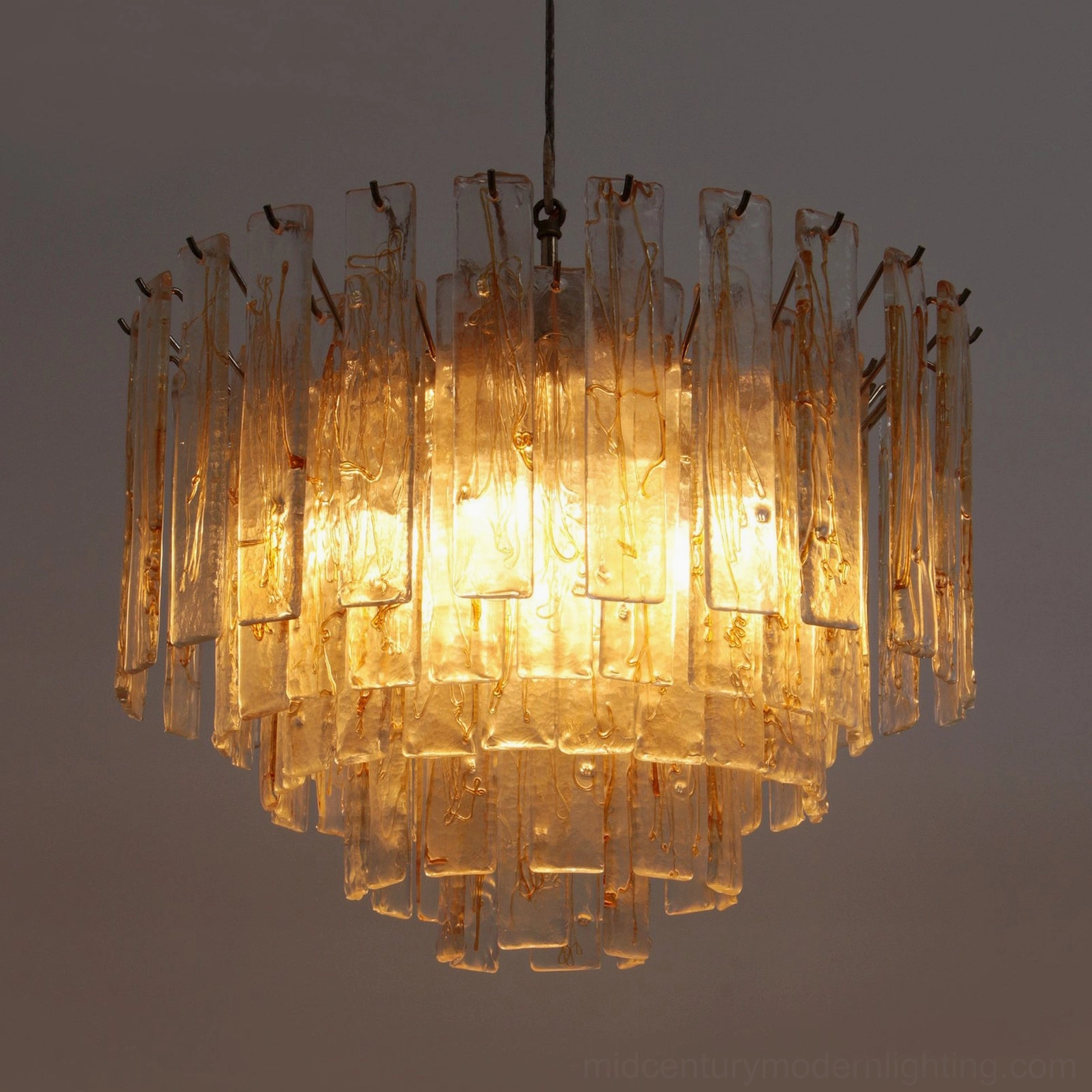 Lit tiered glass chandelier glowing with warm amber illumination against dark backdrop, showing light diffusion through glass