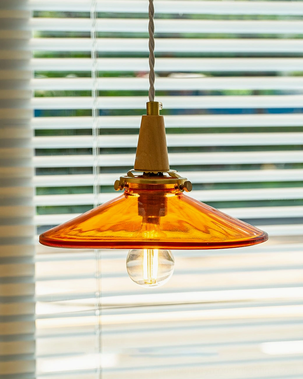 Front-on view of amber glass pendant with wide disc shade, twisted fabric suspension cord, exposed filament bulb against window with horizontal blinds