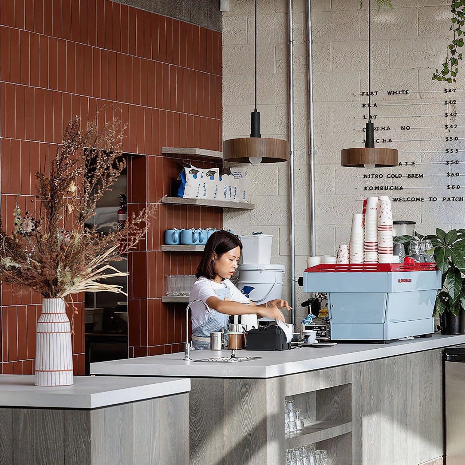 Angilia pendant lights above specialty coffee shop counter with barista, warm wood-tone fixtures complementing retro tiled wall aesthetic
