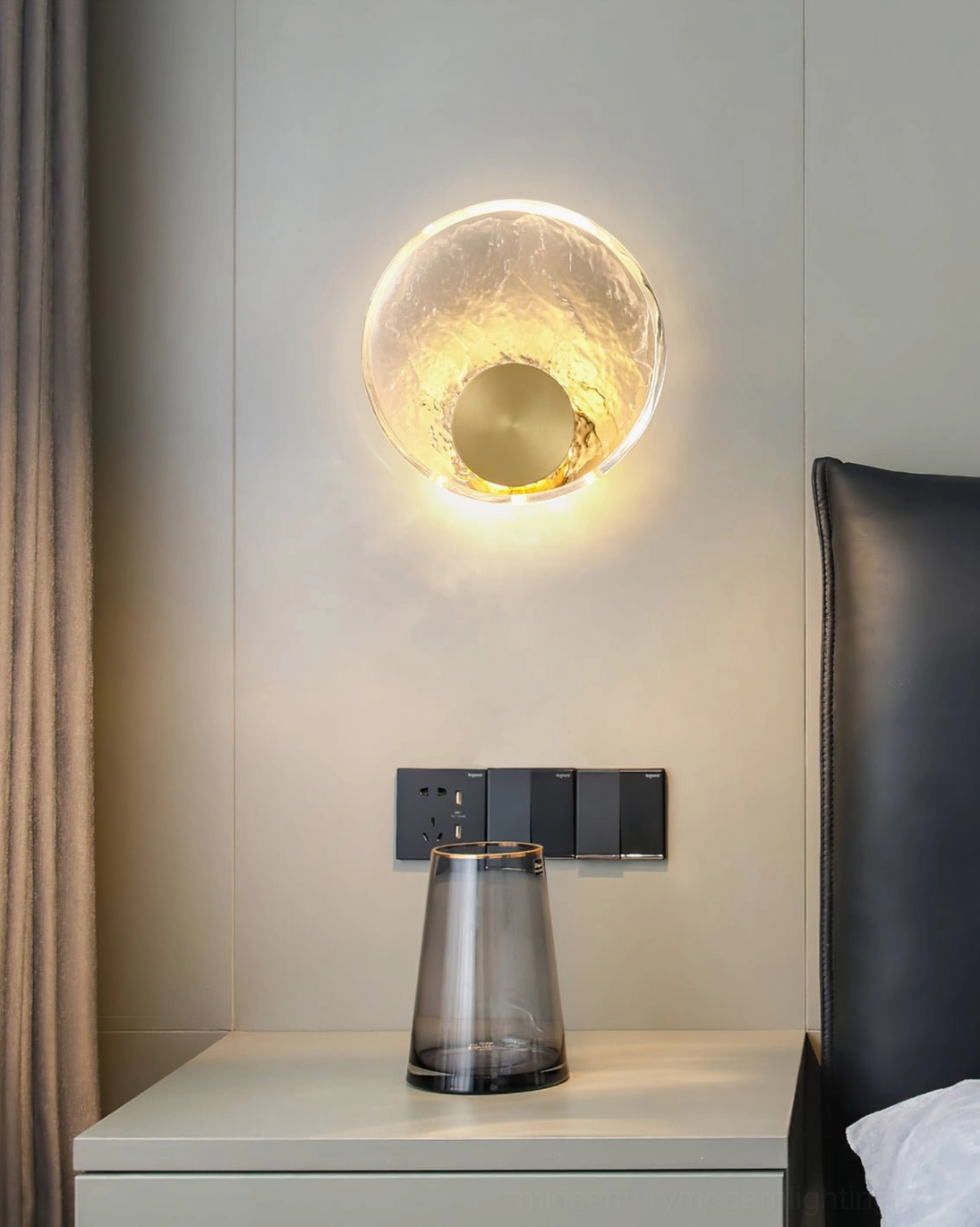 Brass wall sconce above leather headboard in bedroom, crystal shade design, glass bedside accessories, modern mount
