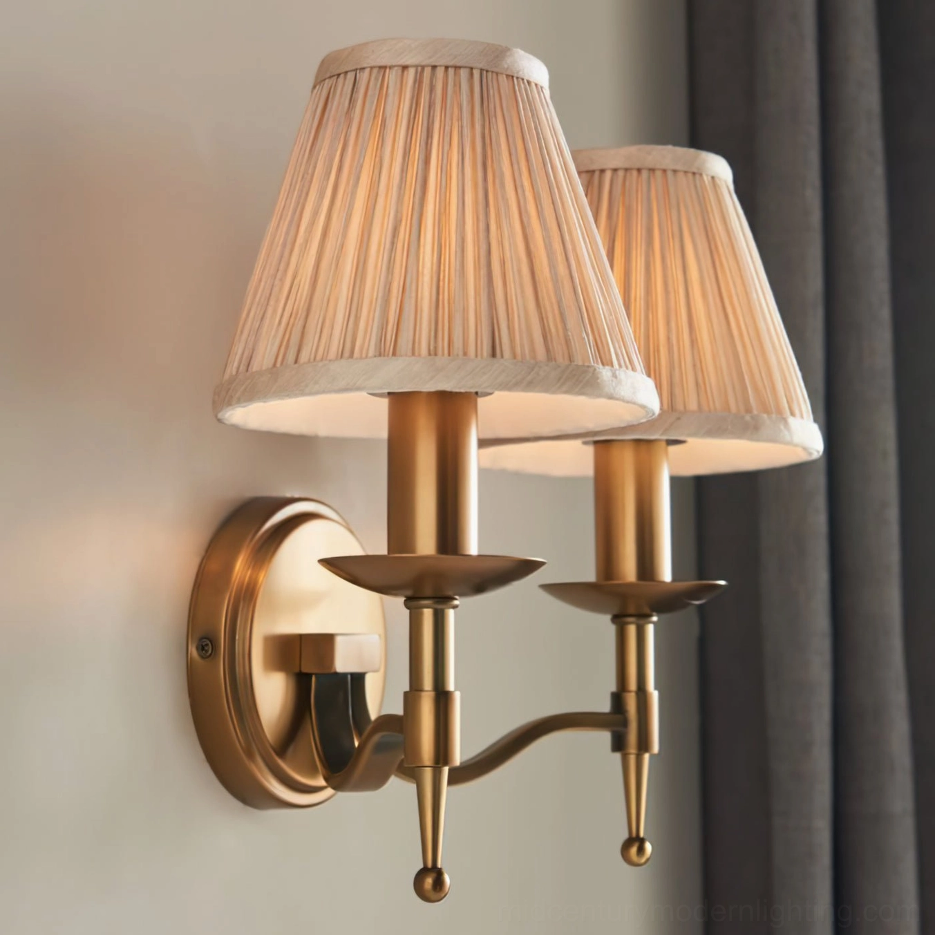 Baylor double-arm brass wall sconce in warm patina finish mounted in modern interior with dark curtain