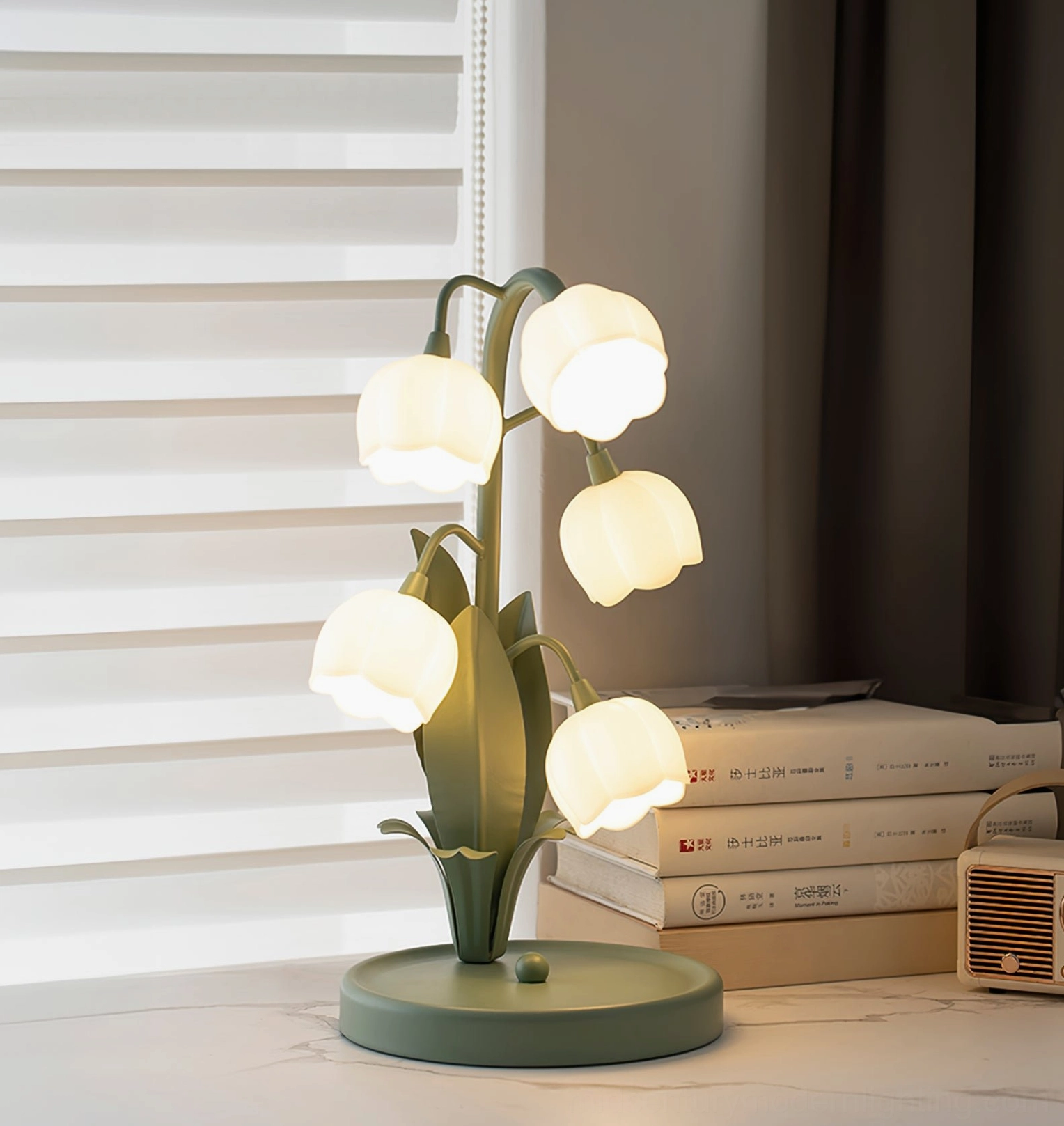 Bell Orchid Table Lamp illuminated on indoor side table with stacked books and window blinds in background