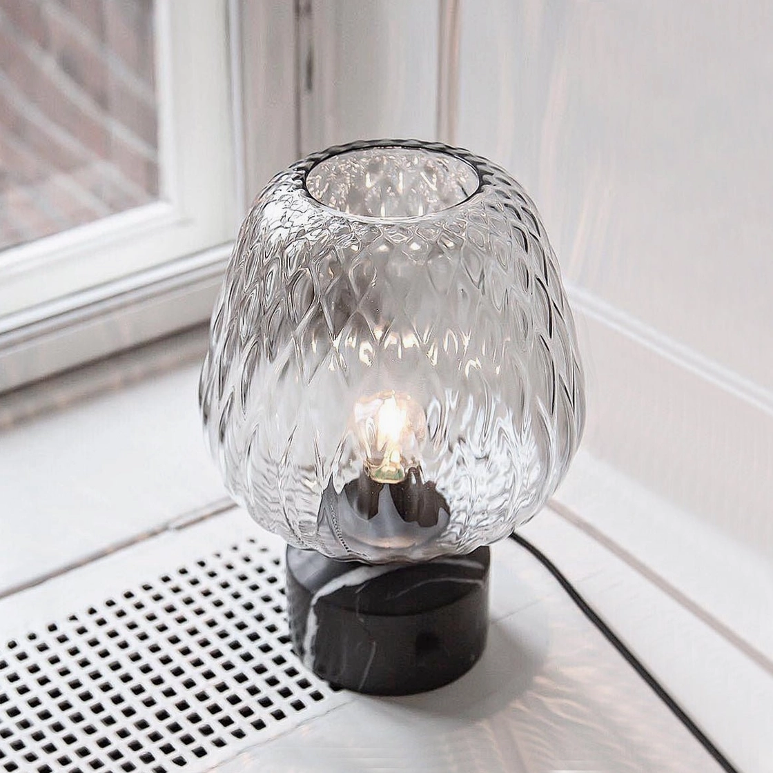 Illuminated blown glass table lamp on windowsill, demonstrating rippled light diffusion and contemporary minimalist aesthetic