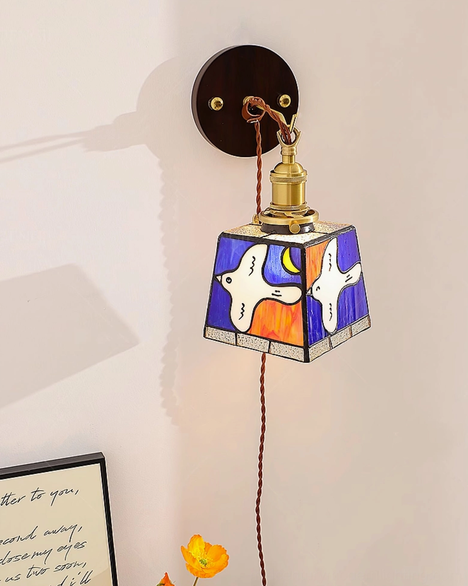 Blue-toned trapezoid stained glass dove wall lamp with brown hanging cord in bedroom setting with floral decor