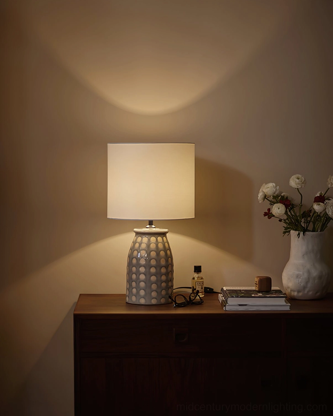 Ceramic patterned table lamp glowing on dark wooden dresser with flower vase and decorative stacked items