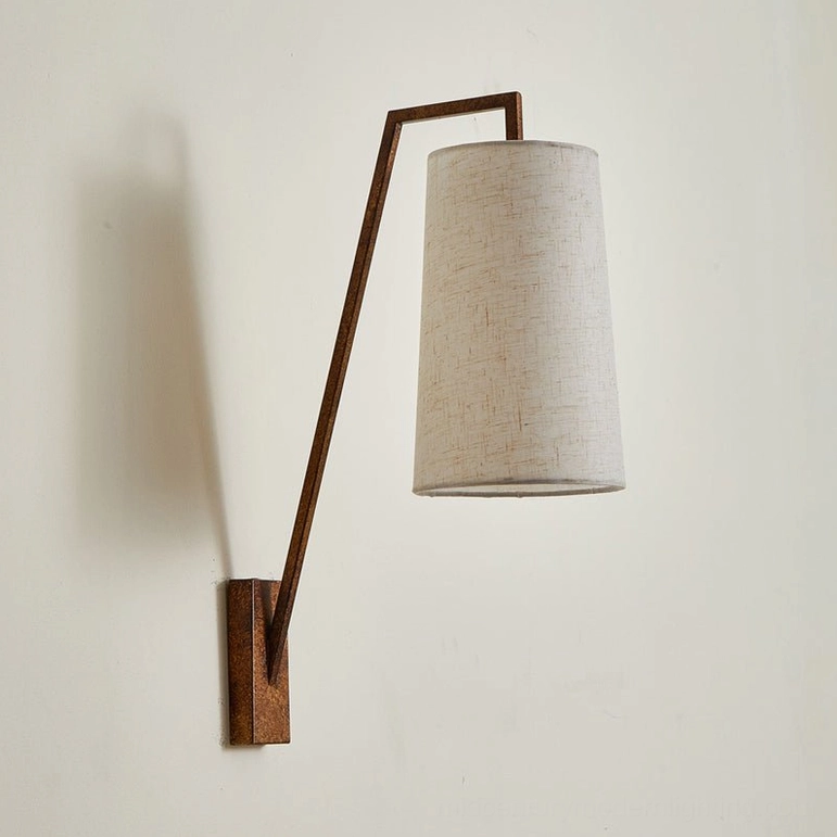 Antique bronze Bonny wall sconce with weathered metal arm and beige fabric shade, mid-century modern design on light wall