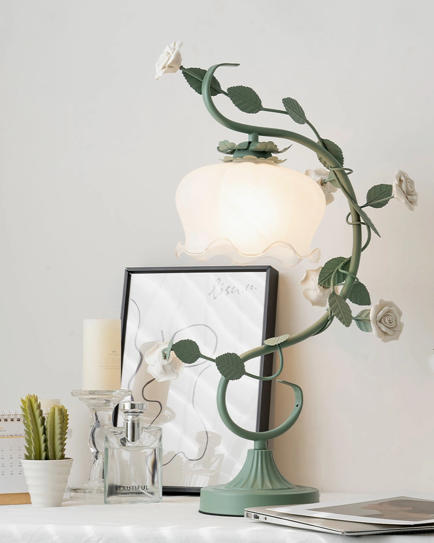 Unlit rose vine table lamp oblique view with warm frosted glass shade, candle and art frame nearby