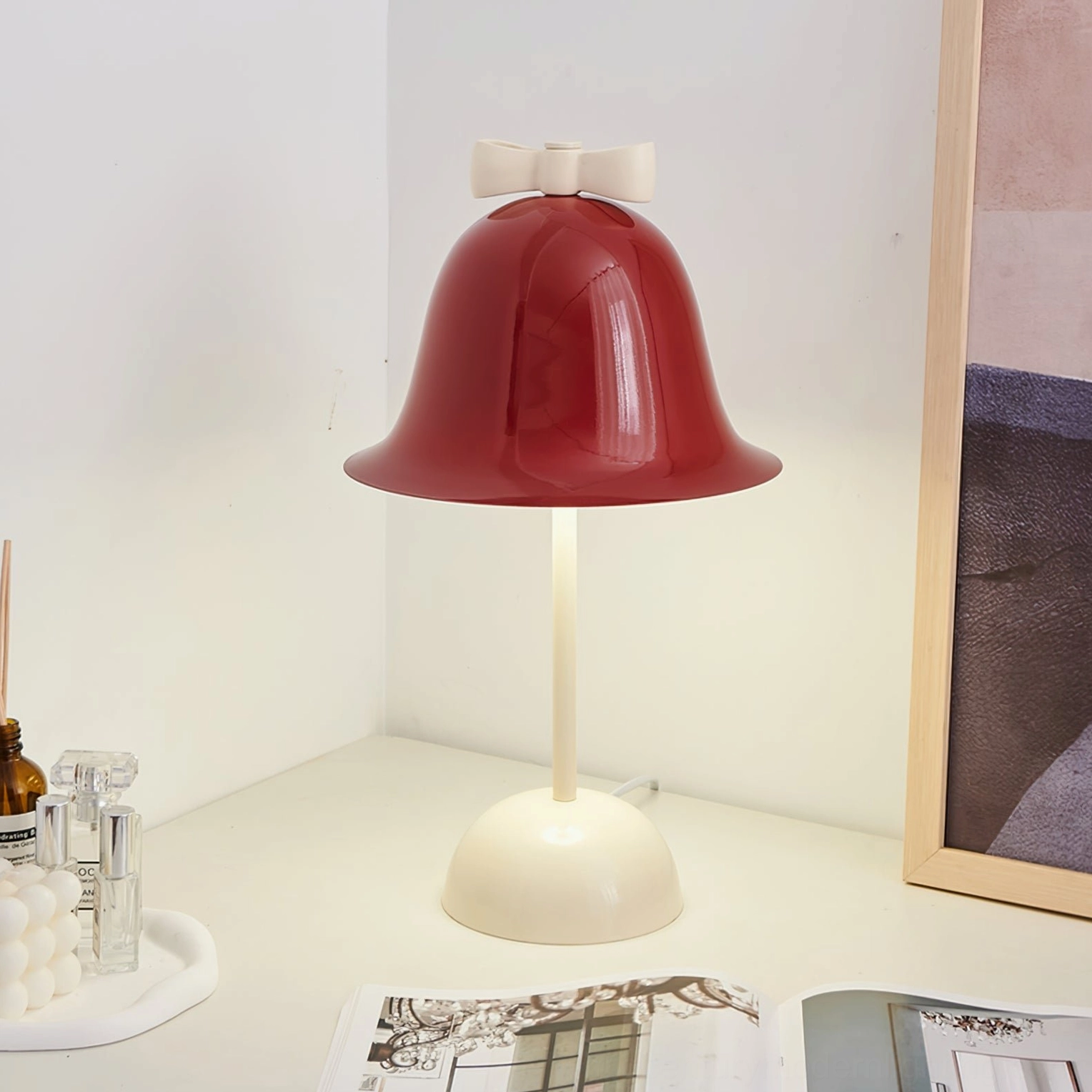 Red burgundy Bow Bell table lamp on light-colored side table with skincare bottles, magazines, and framed art print decor