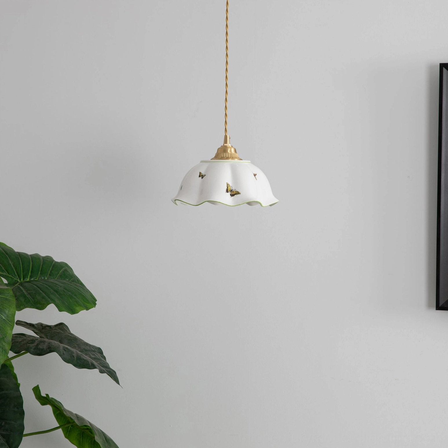 Unlit ceramic butterfly pendant with scalloped edge shade displayed against plain wall with framed artwork
