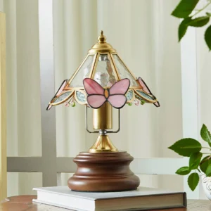 Butterfly table lamp with brass hardware displayed on wooden side table with books and green plants, three-quarter view