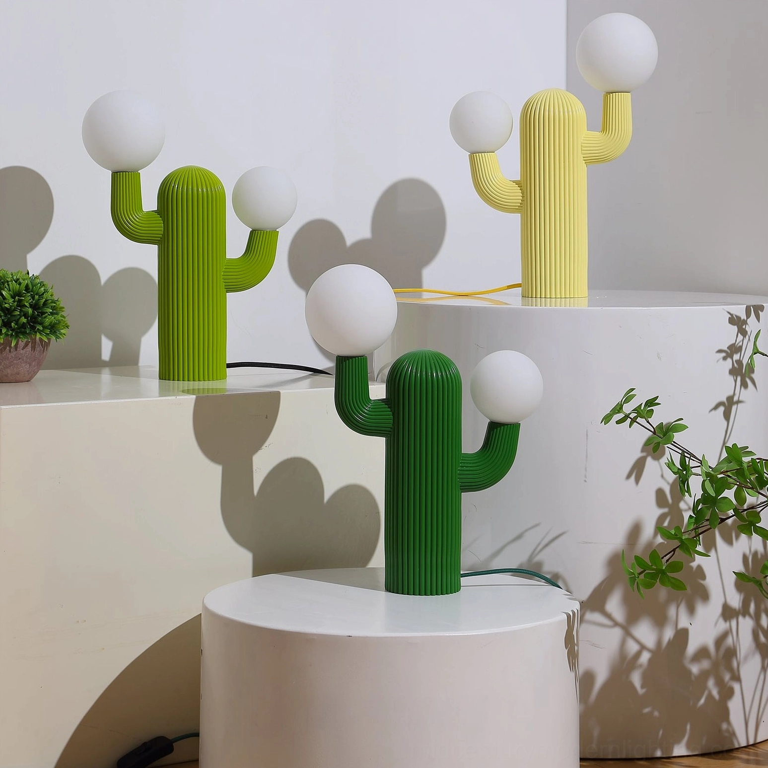 Multiple cactus table lamps in green and yellow finishes displayed on white pedestals, showing color variations and sculptural form