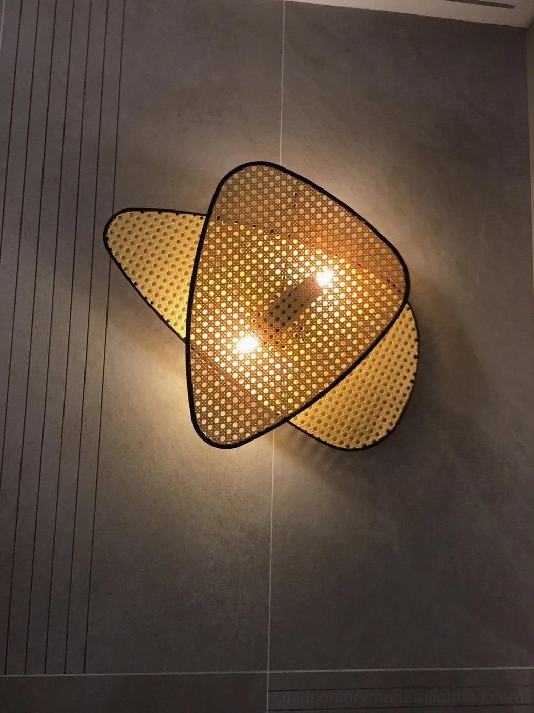 Woven rattan wall lamp illuminated on dark grey striped tiled wall showing layered cannage pattern and warm glow