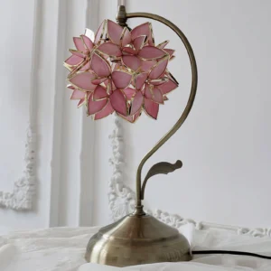 Pink capiz shell lamp displayed on ornate bedroom bedside table, three-quarter angle showing curved bronze metal arm and petal details