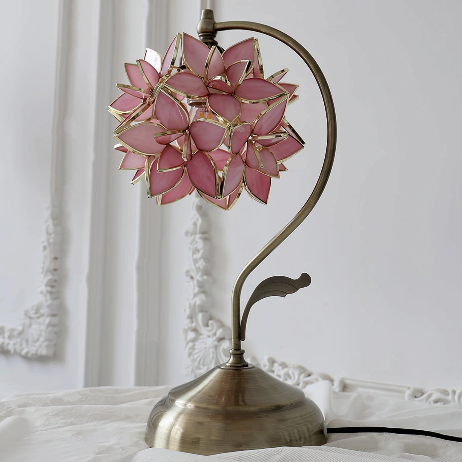 Pink capiz shell lamp displayed on ornate bedroom bedside table, three-quarter angle showing curved bronze metal arm and petal details
