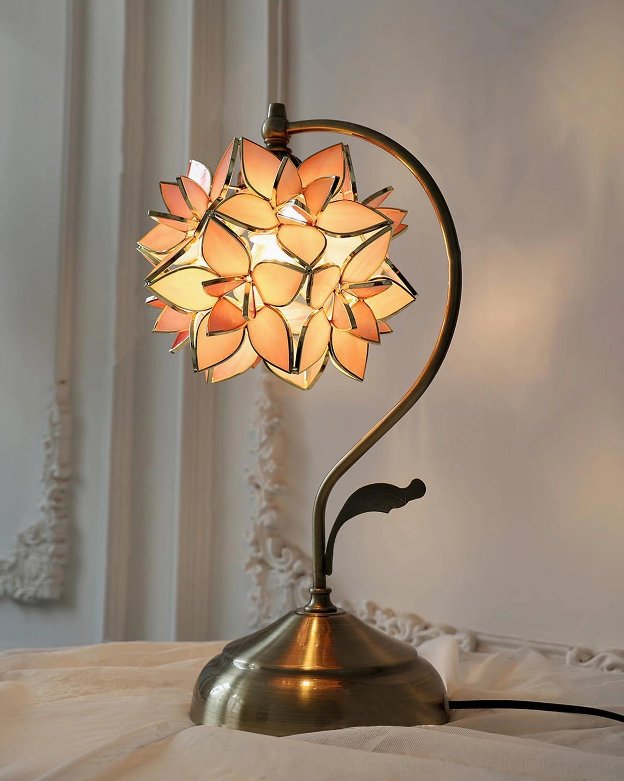 Lit orange capiz flower lamp illuminated on bedroom nightstand, warm amber glow through stained glass petals, three-quarter view