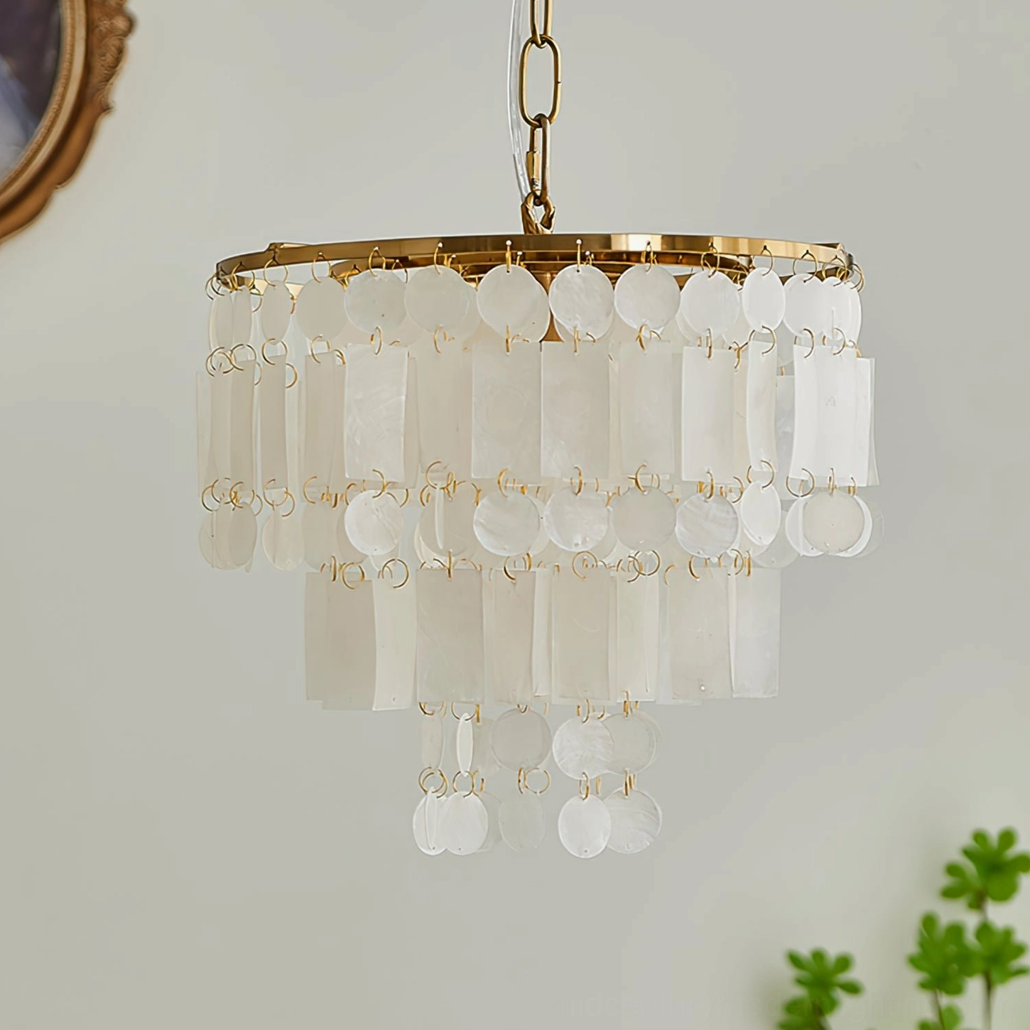 Coastal luxury capiz shell chandelier with translucent white shell pendants and brass gold frame, surrounded by green plants