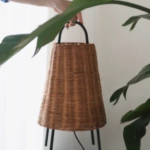 Hand holding Catalina Coastal Table Lamp with woven rattan shade and black metal frame, indoor with green foliage backdrop