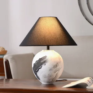 Ceramic table lamp with lit shade creating warm glow on wooden desk with open book and interior styling