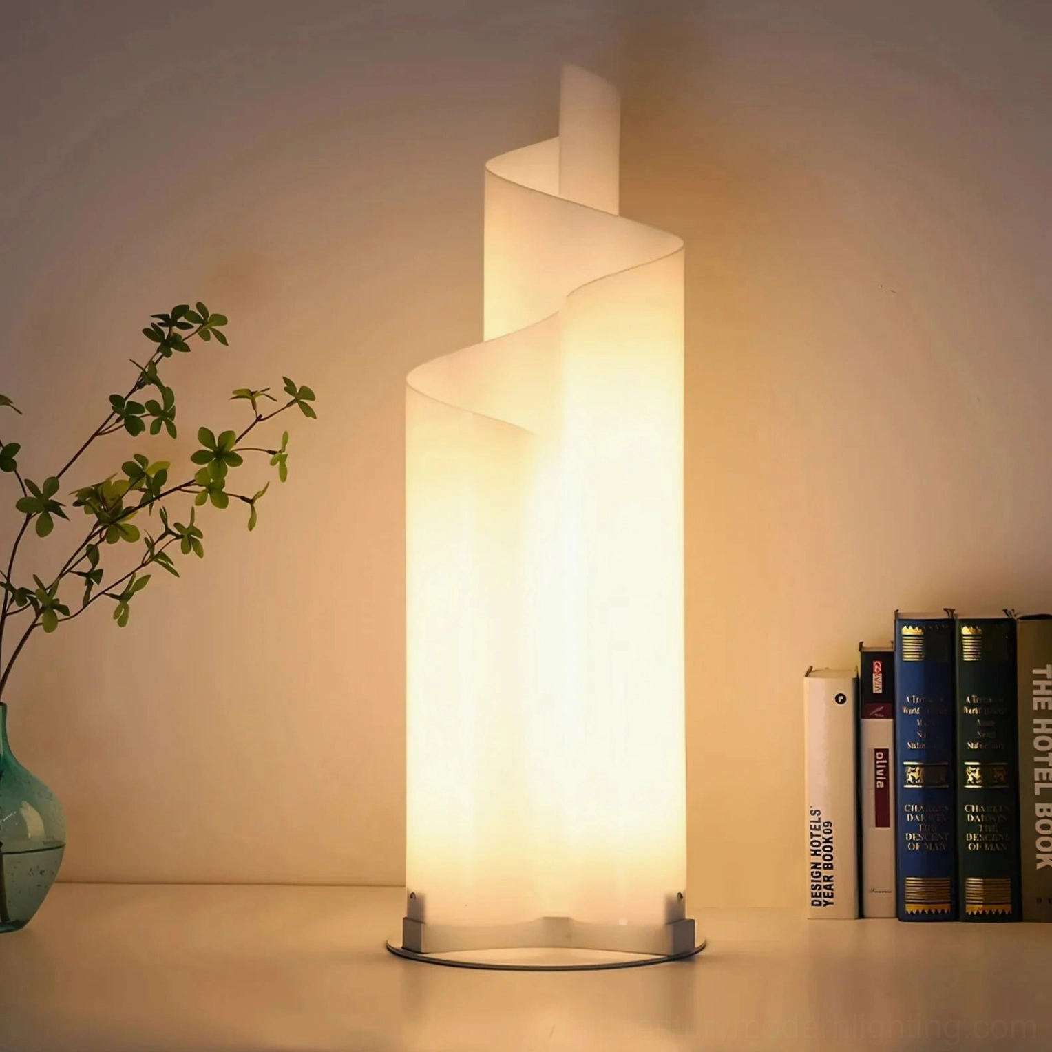 Warm glowing chimera spiral table lamp three-quarter view against warm wall with greenery vase and book stack styling