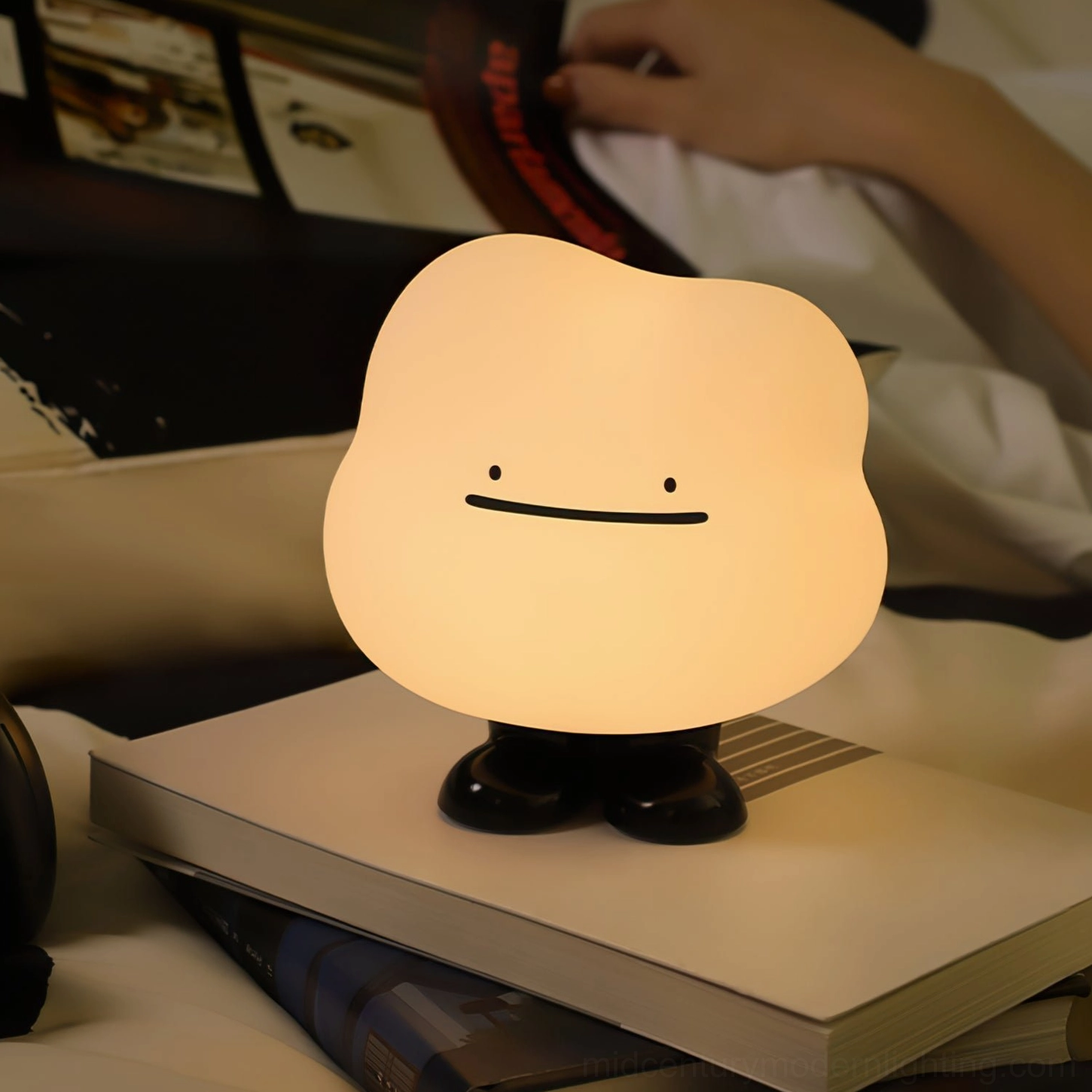 Cloud Night bedside lamp placed on stacked books providing warm ambient lighting for person reading on bed