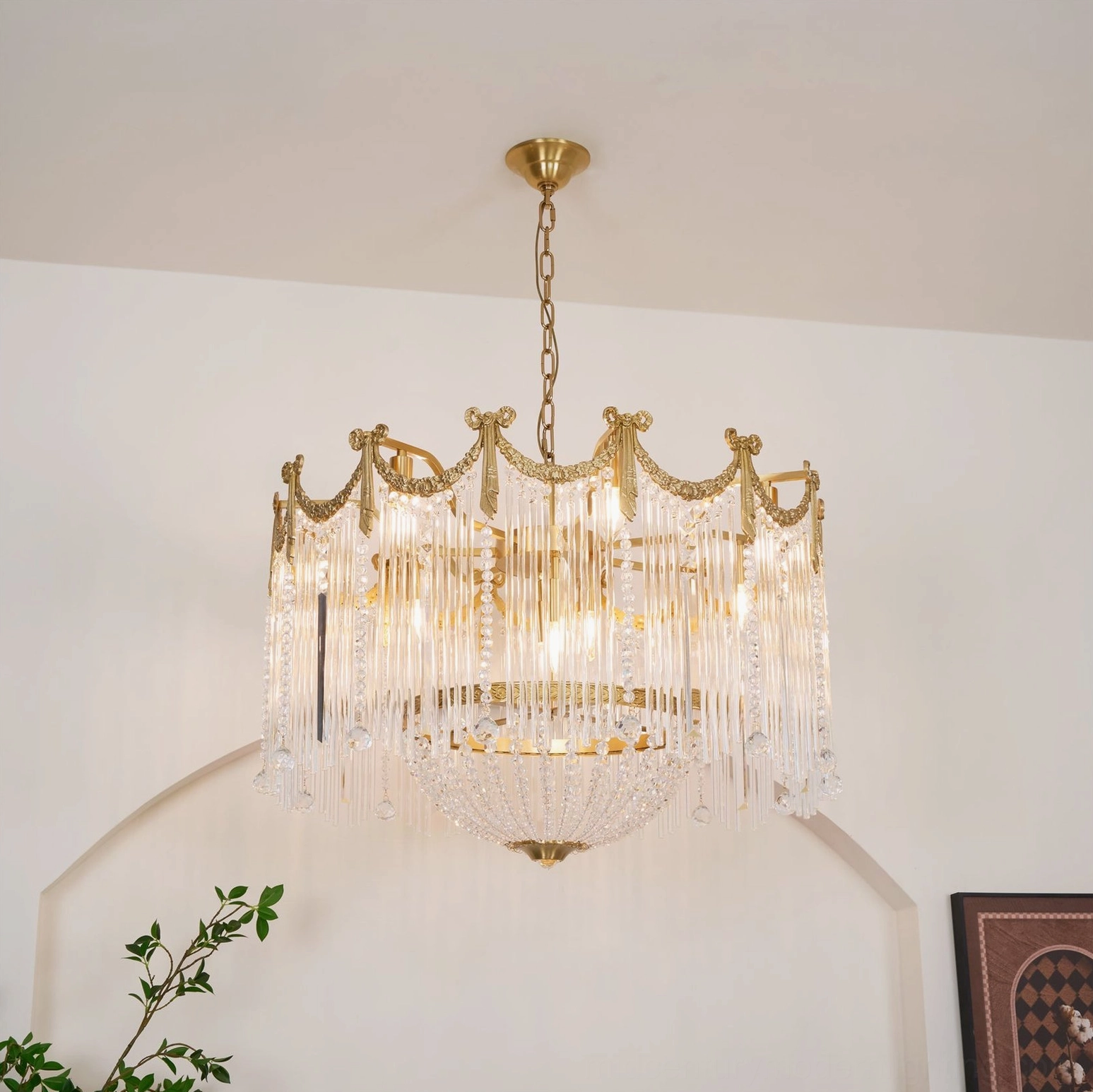 Three-quarter view of crystal teardrop chandelier suspended by metal chain with crown frame against white wall and plants