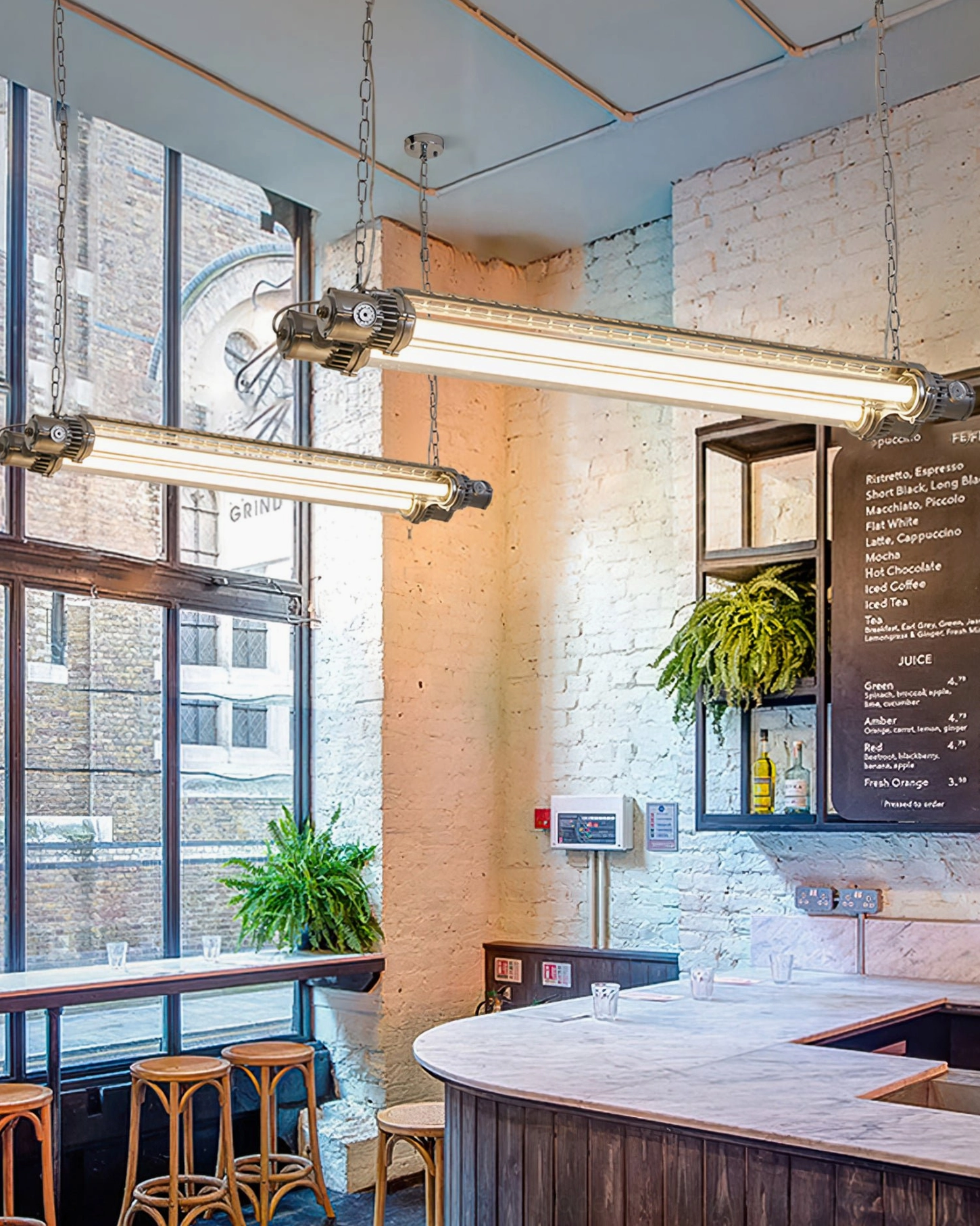 Dual industrial silver tube chandeliers hanging above brick wall coffee shop counter, matching pendant fixtures