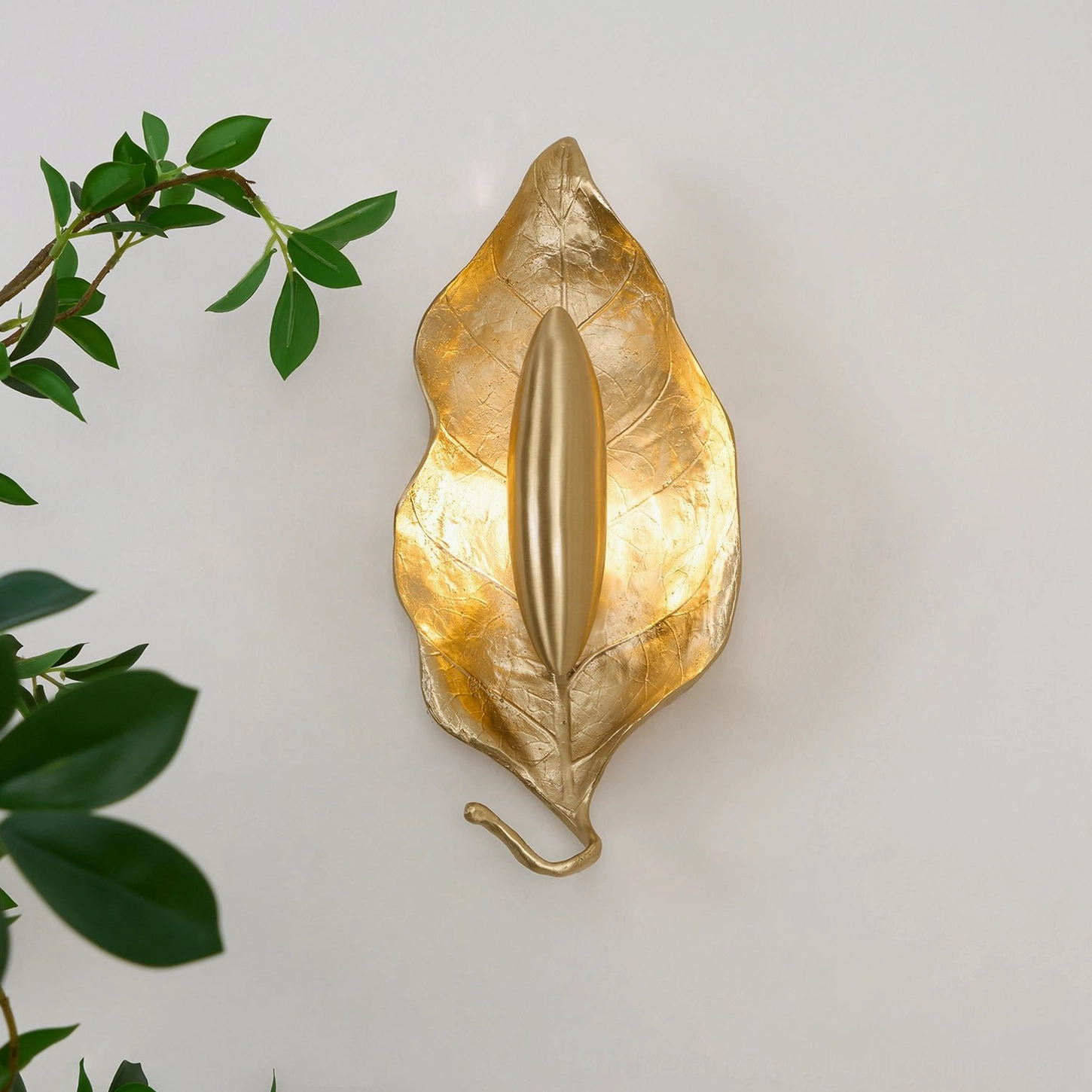 Dumaine brass leaf wall lamp mounted on white wall with green branches, showing realistic leaf venation texture under light