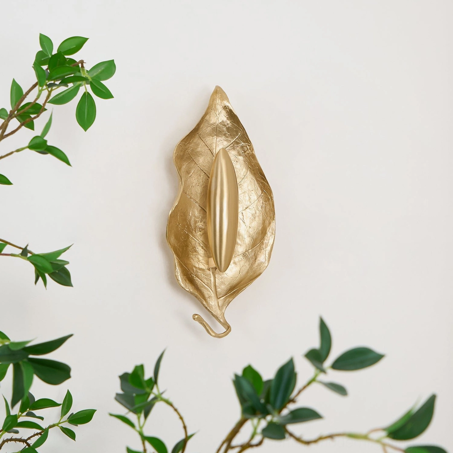 Compact Dumaine brass leaf wall lamp top-down angle against white wall with scattered green foliage accents