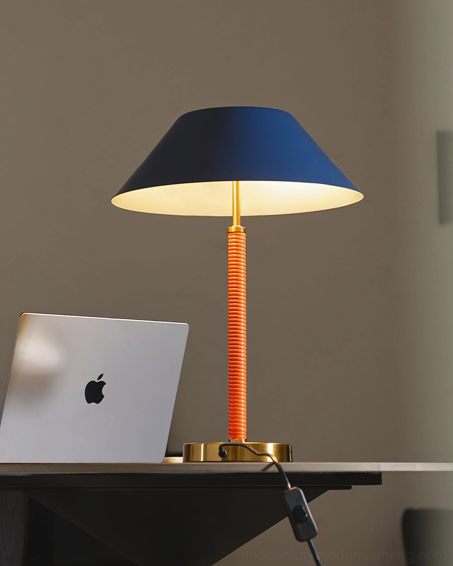 Eave table lamp illuminated on minimalist desk setup, showing warm glow from interior navy shade and brass gold base