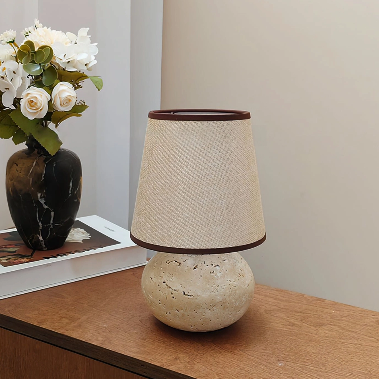 Unlit Elba table lamp with linen shade dark brown trim on wooden bedside table, left three-quarter perspective of sculptural travertine base