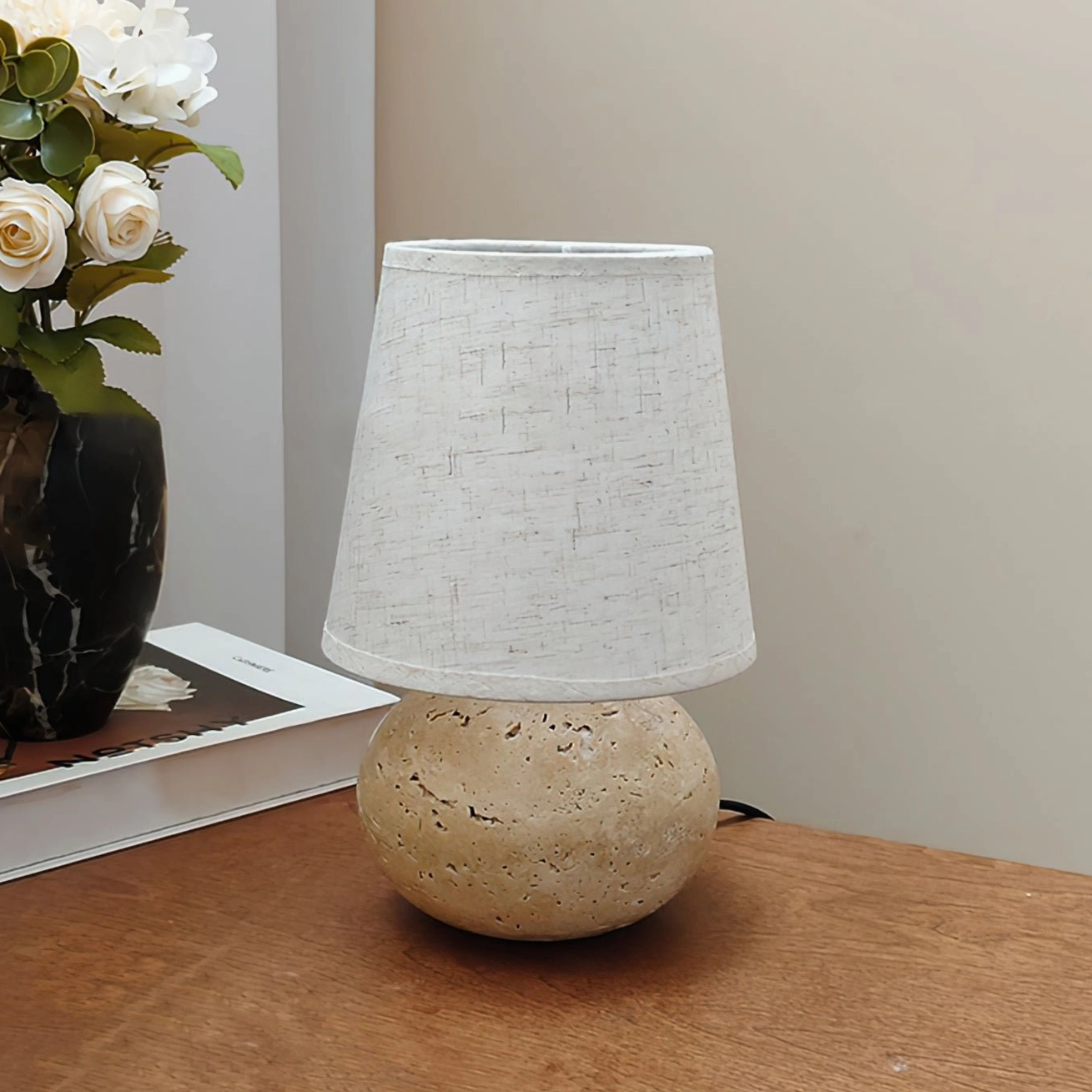 Elba table lamp with white linen shade no trim left three-quarter front view on wooden bedside table, unlit travertine base