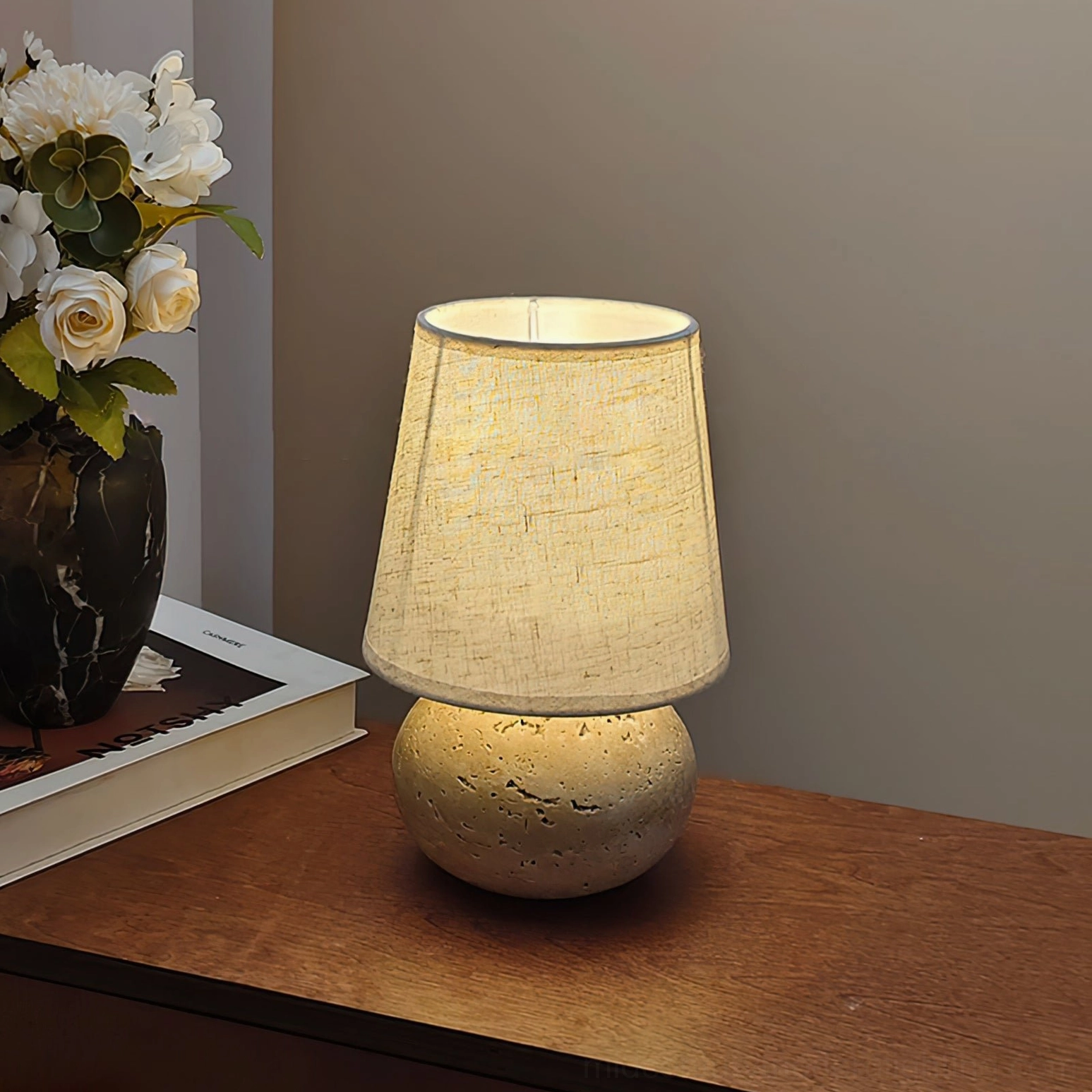 Lit Elba travertine table lamp on wooden bedside with neutral warm styling, front view showing plain linen shade and warm ambient light