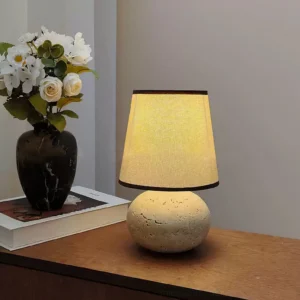Lit Elba travertine table lamp on wooden bedside table, ambient warm light with marble vase and stacked books, three-quarter view