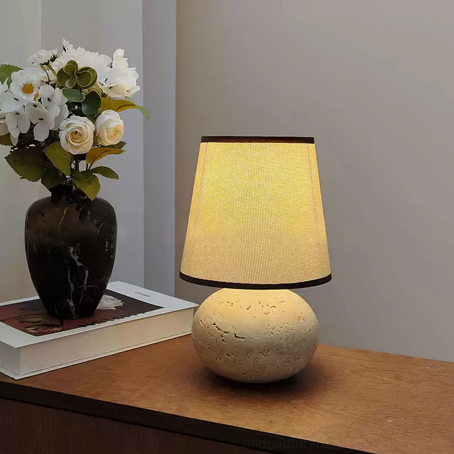 Lit Elba travertine table lamp on wooden bedside table, ambient warm light with marble vase and stacked books, three-quarter view