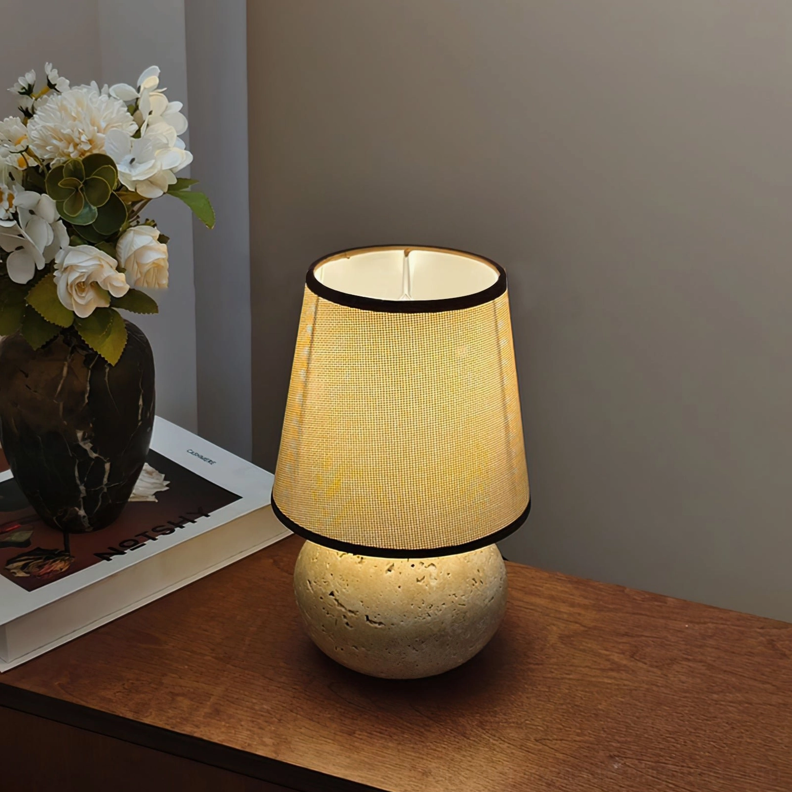 Side three-quarter view of glowing Elba travertine lamp on wooden bedside, close-up of rustic stone base and warm light diffusion