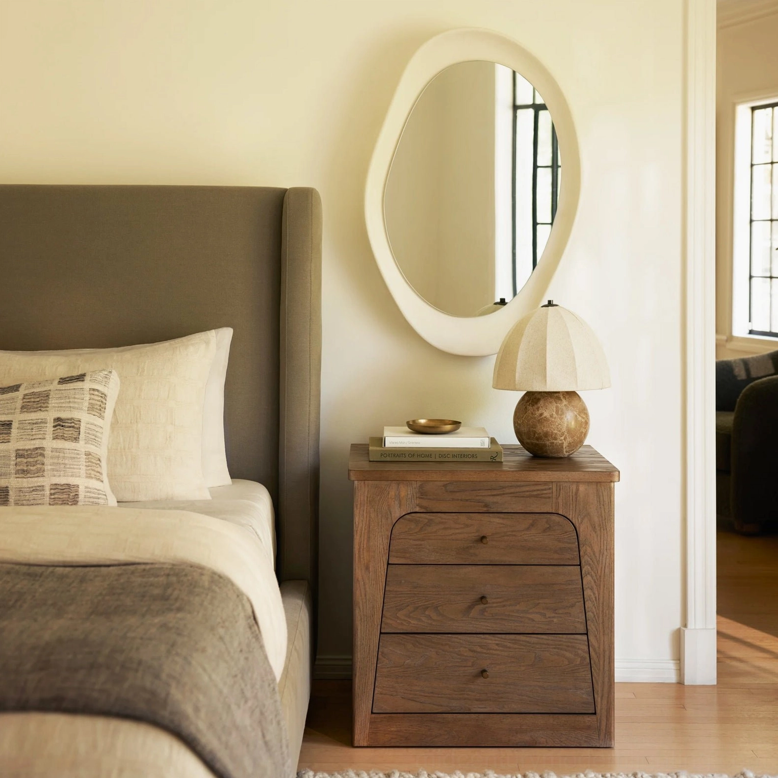 Enora organic modern lamp on wooden nightstand with oval mirror, warm neutral bedroom palette, compact modern design