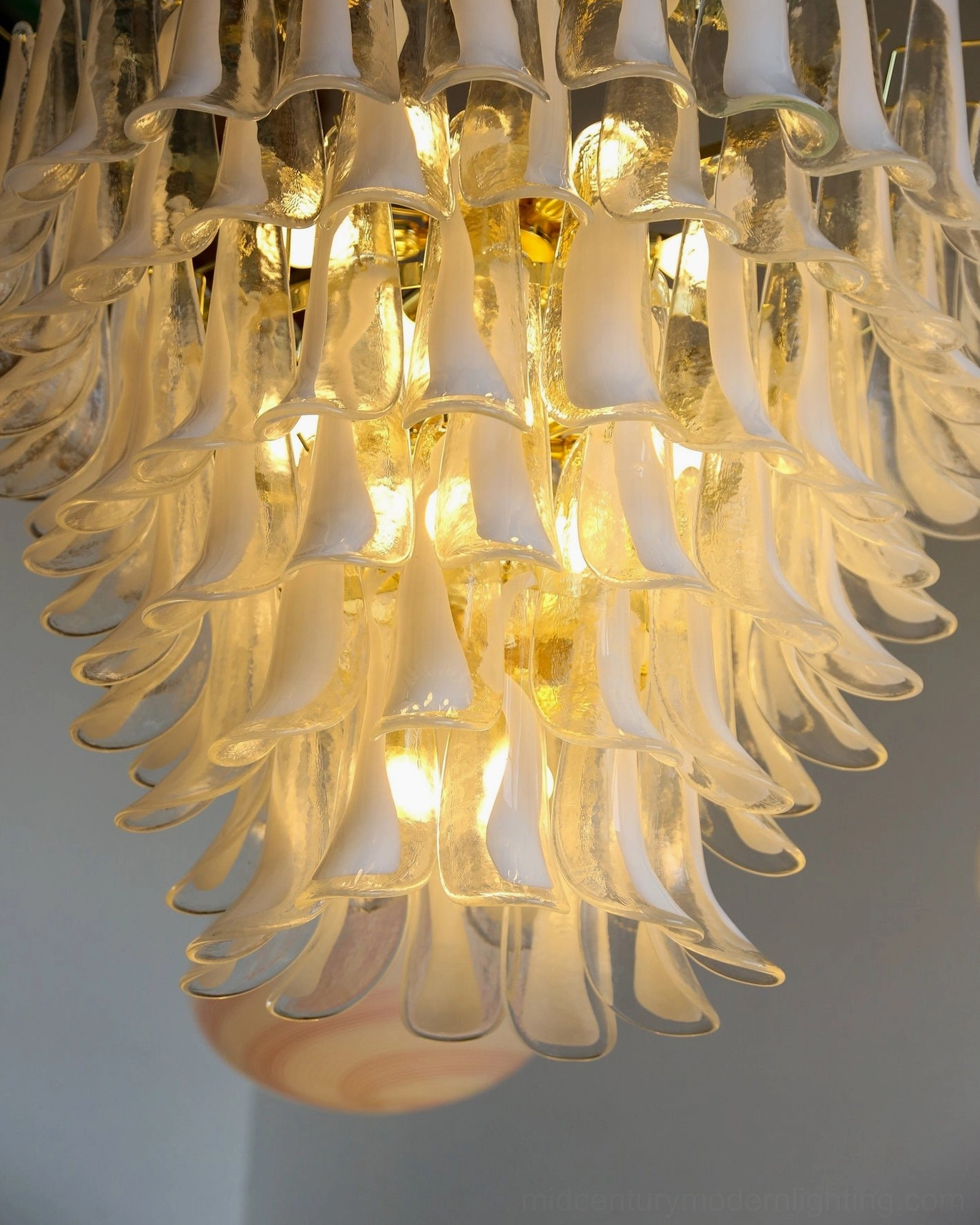 Low-angle close-up of glowing petal chandelier showing warm golden light distribution through layered clear glass petals against wood trim wall
