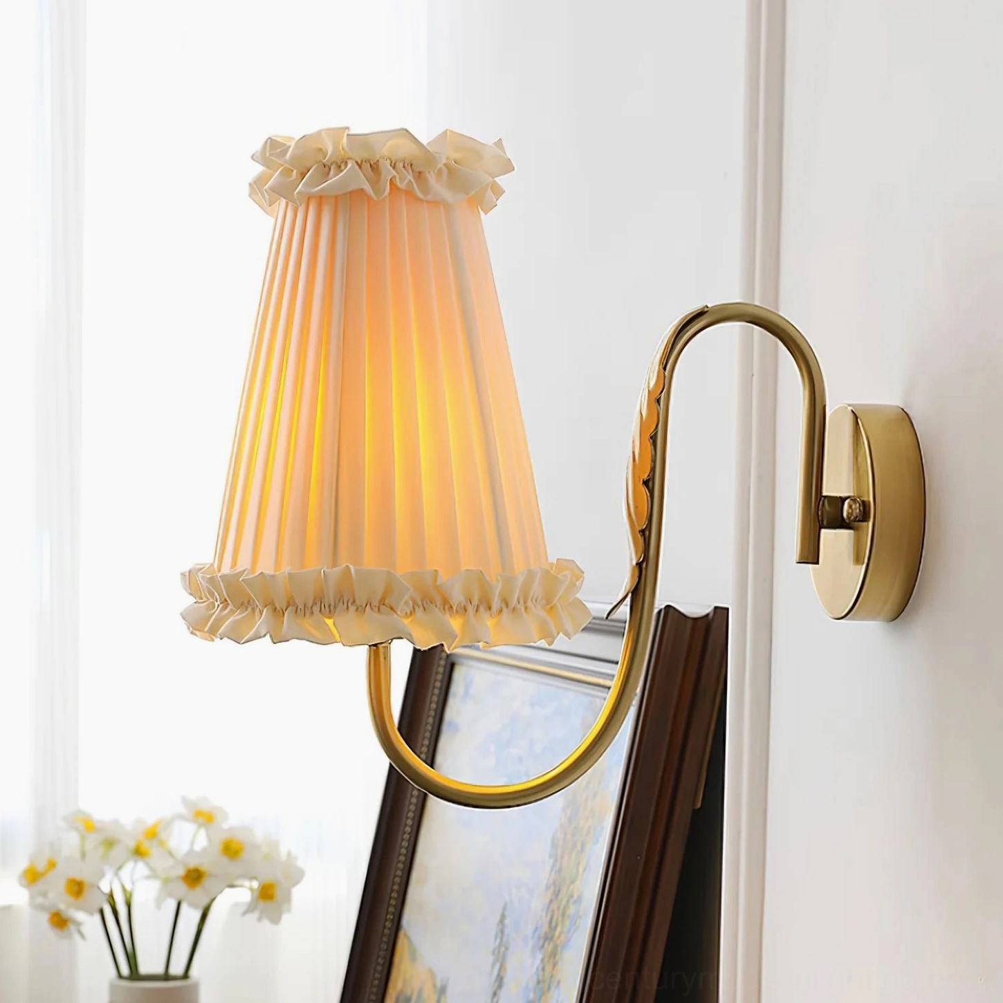 Wall-mounted brass sconce with beige pleated shade positioned above flowers in home decor setting
