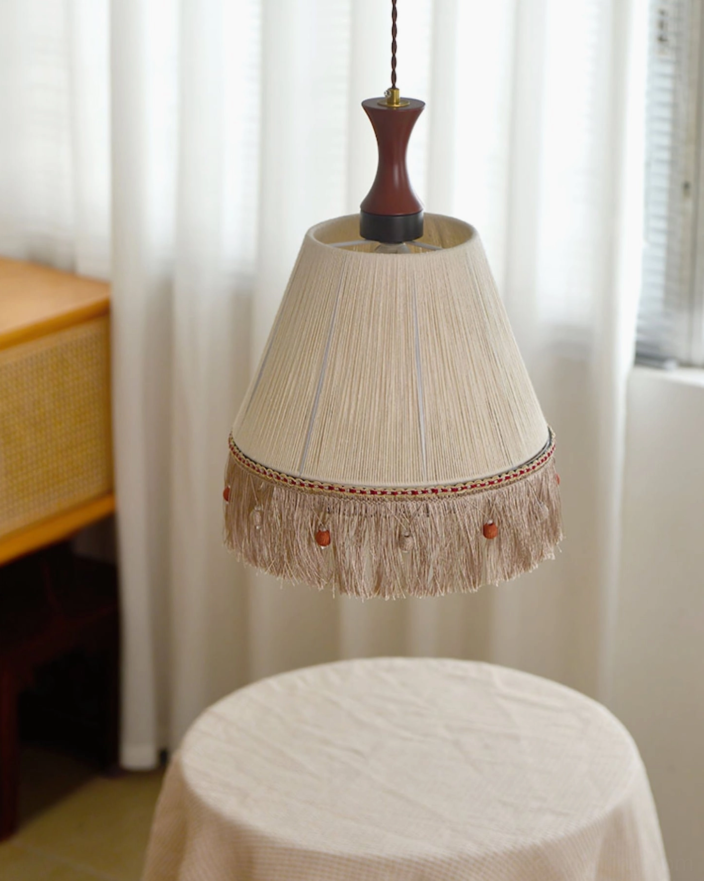 Beige fringe tassel pendant lamp glowing warmly above round dining table with sheer curtains