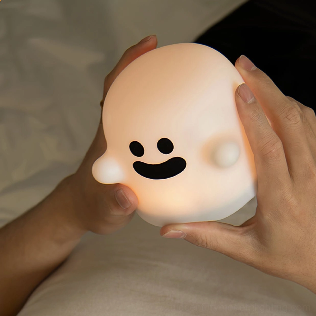 Ghost night light with happy expression being gently held in two hands showing soft silicone material