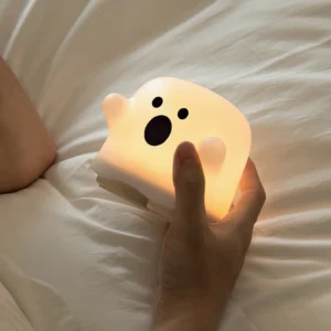 Ghost silicone night light with warm glow in bedroom setting showing cute expressive face held above linen bedding