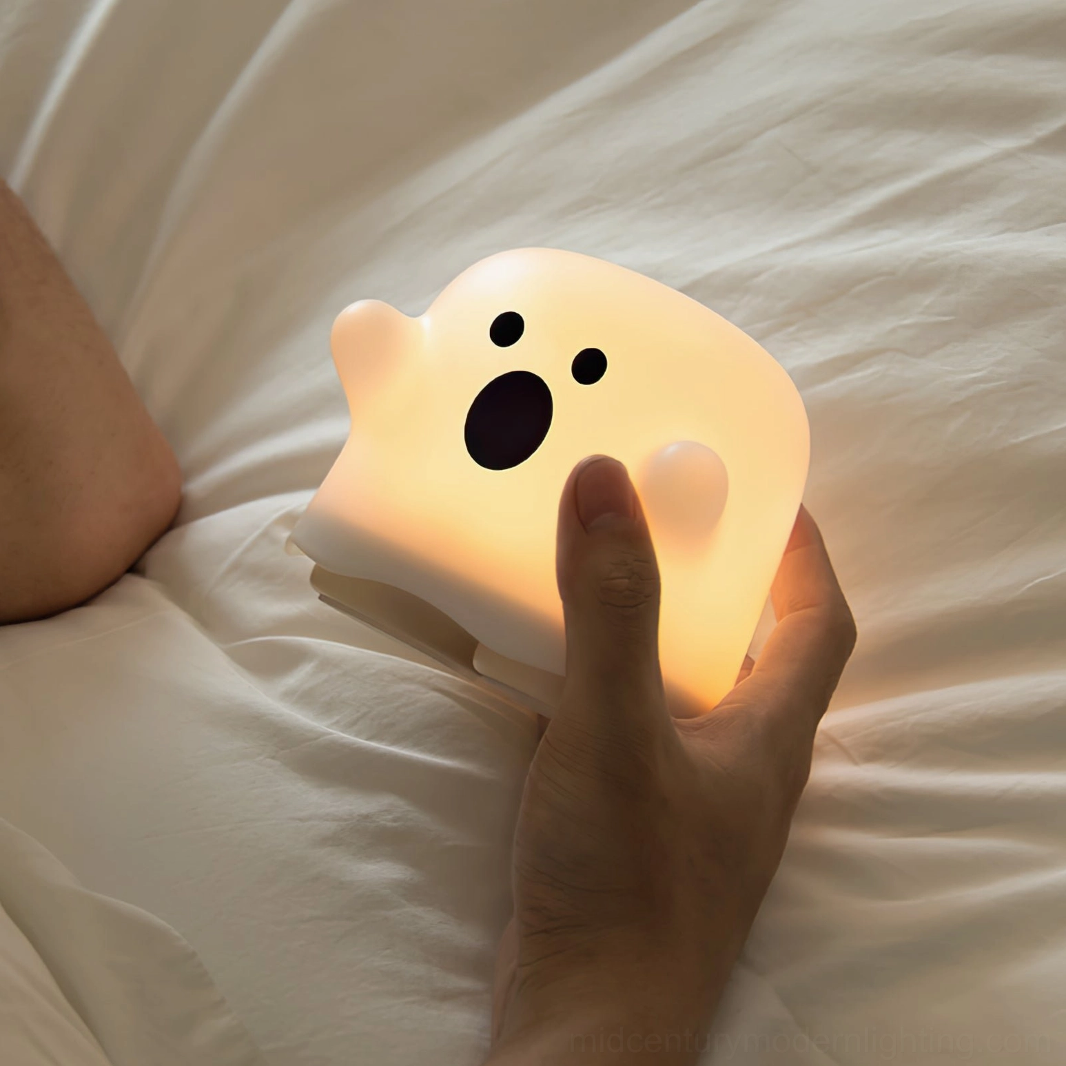 Ghost silicone night light with warm glow in bedroom setting showing cute expressive face held above linen bedding