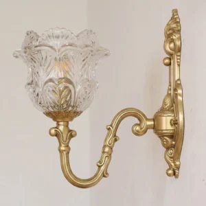 Close-up detail of Gideon wall sconce showing ornate brass base with embossed detailing and clear tulip glass lampshade on light gray wall