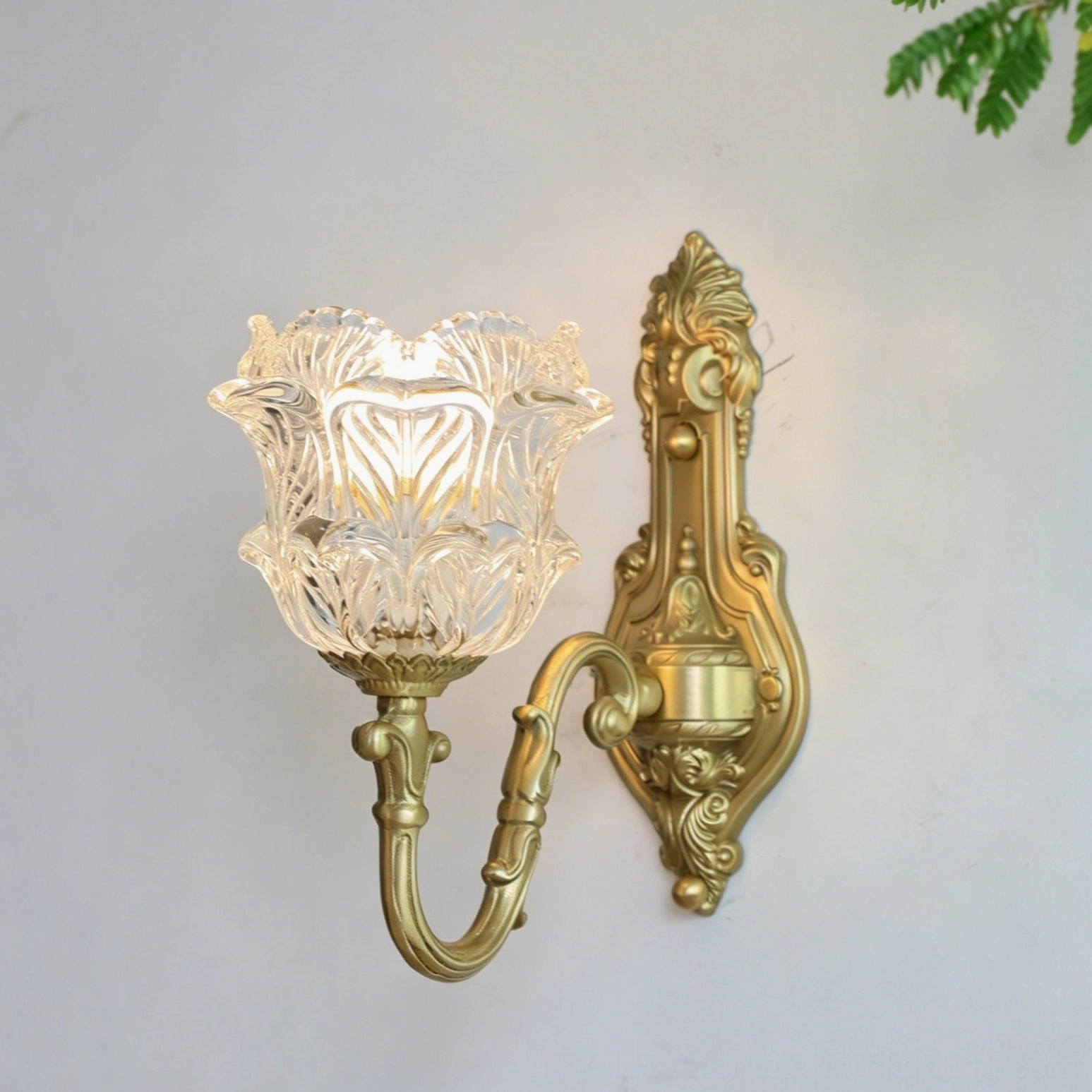 Illuminated Gideon wall sconce photographed against light gray background, showcasing warm glow through clear tulip glass and brushed brass frame