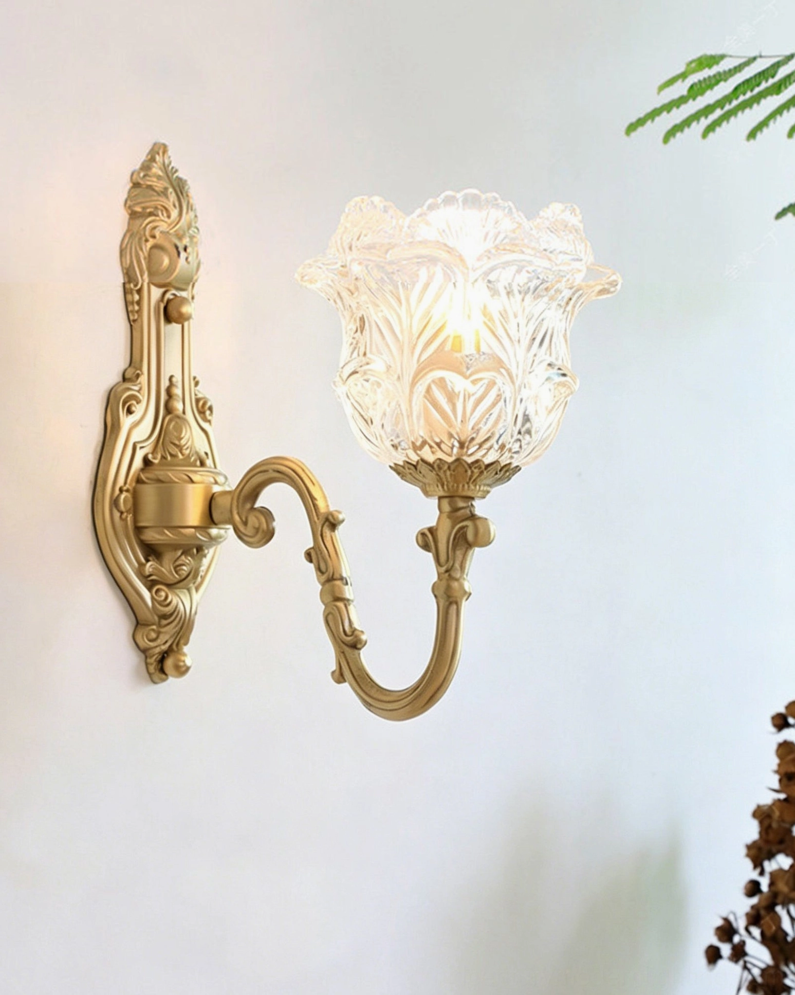 Illuminated Gideon Baroque wall sconce mounted on indoor wall with warm light glowing through tulip glass shade, featuring carved brass scroll arm