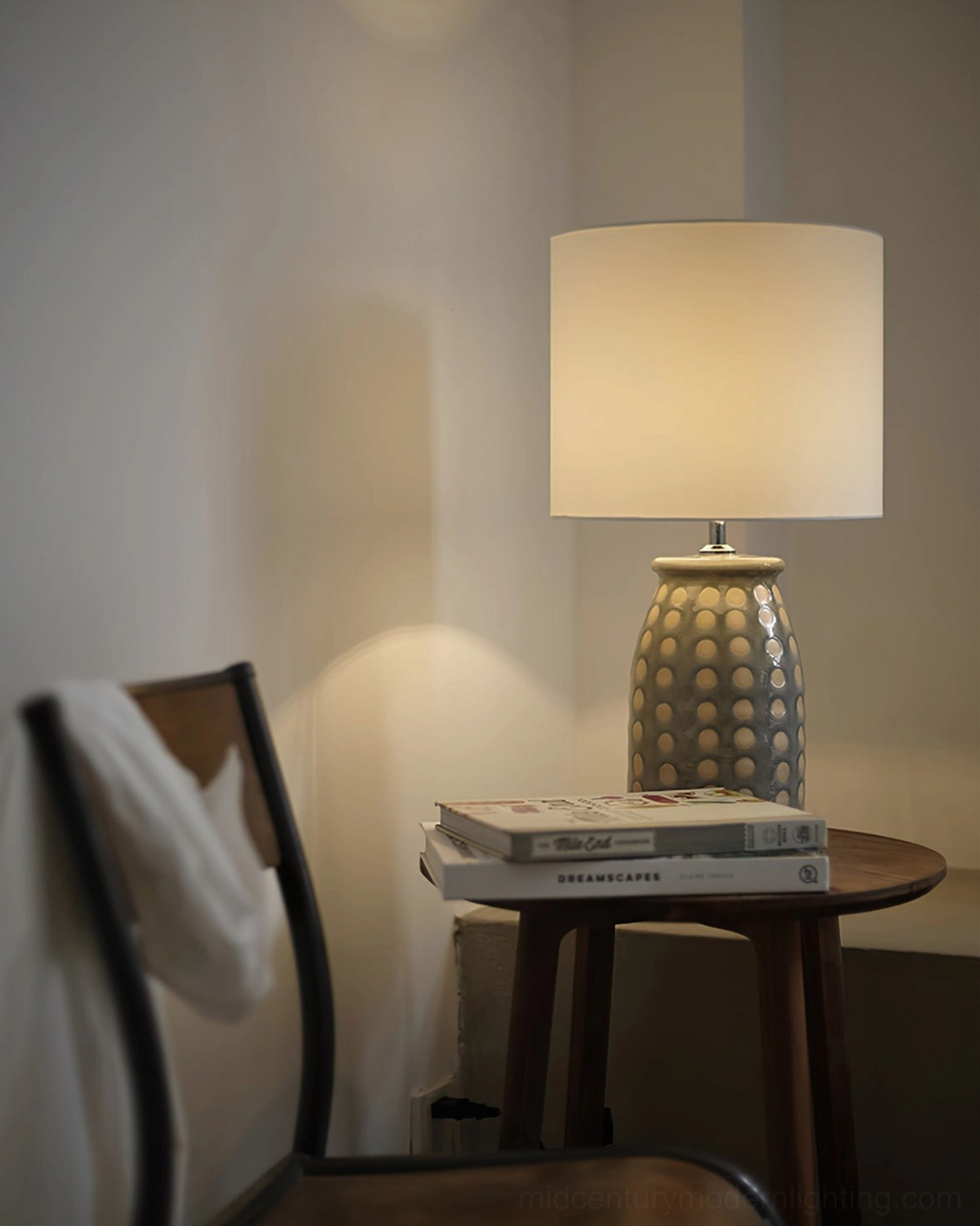 Lit ceramic patterned lamp on wooden side table with draped white chair, warm ambient lighting in modern interior