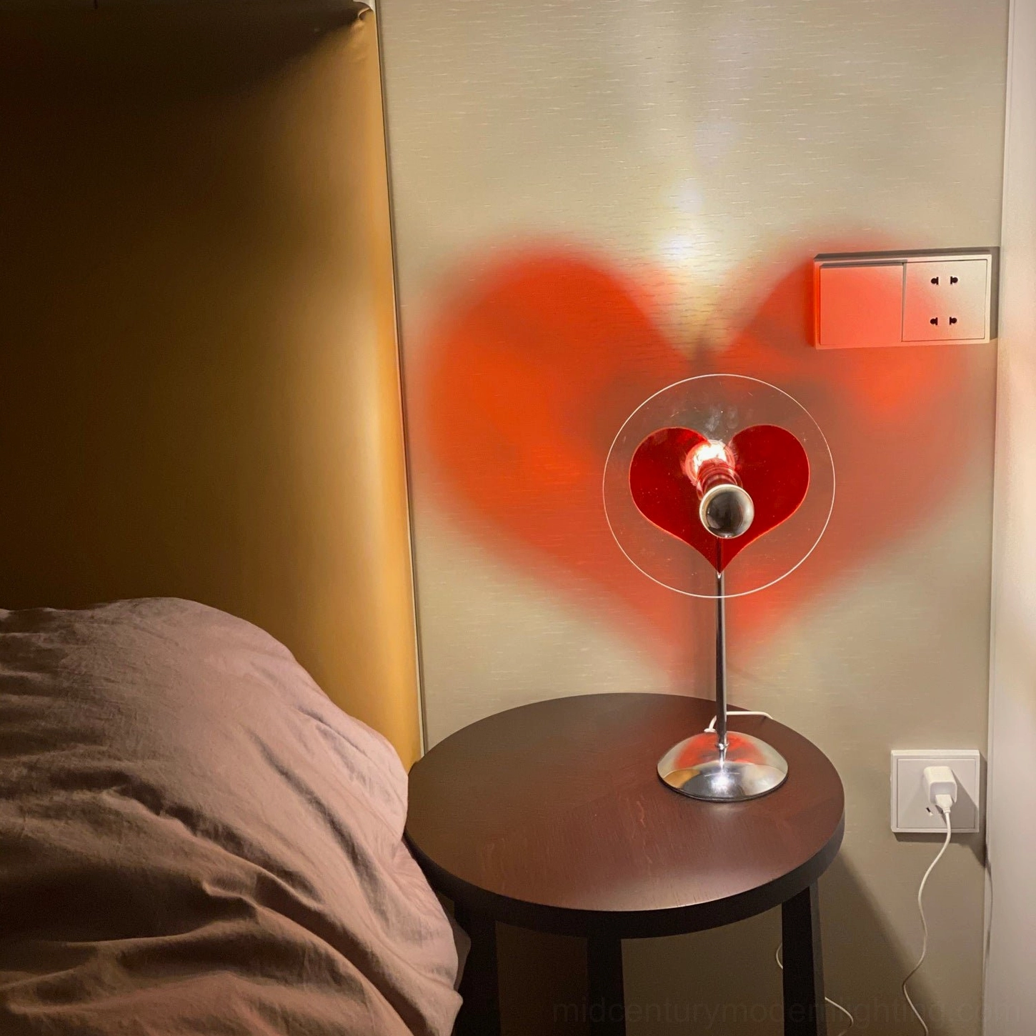 Heart projection lamp on wooden nightstand, casting warm orange-red heart silhouette on bedroom wall